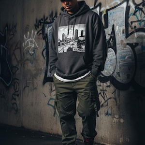 May include: A charcoal grey hooded sweatshirt featuring a black and white city skyline graphic print on the chest, paired with olive green cargo trousers, a camouflage baseball cap with a red logo patch, and black and red high-top trainers.