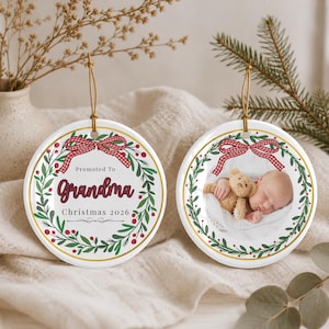 Può includere: Due ornamenti rotondi in ceramica bianca con bordi dorati e cordini dorati. Uno presenta una ghirlanda verde, bacche rosse, un fiocco a quadretti rosso e il testo Promoted To Grandma Christmas 2026. L'altro mostra un bambino con un orsacchiotto.
