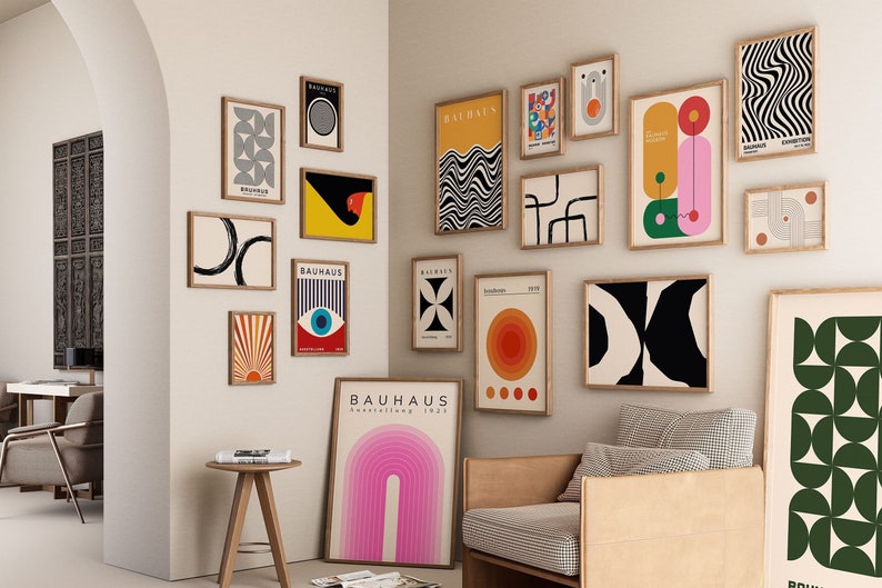 May include: A gallery wall featuring a collection of abstract art prints in various colours and patterns. The prints are framed in natural wood and arranged in a grid pattern. The prints feature geometric shapes, lines, and colours, including black, white, red, yellow, green, and blue. The prints are hung on a white wall.