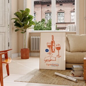 May include: A beige couch with a throw pillow and a stack of books on a wooden table. A framed illustration of a wine bottle and two glasses with the text "Liquid Therapy" is on the wall. A potted plant is in the corner of the room.