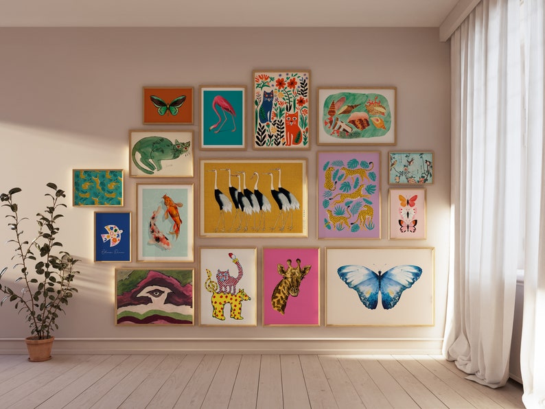 May include: A collection of framed art prints featuring various animal and nature-themed illustrations. The artwork includes images of butterflies, cats, flamingos, koi fish, and giraffes. The prints are displayed on a neutral wall with a potted plant.