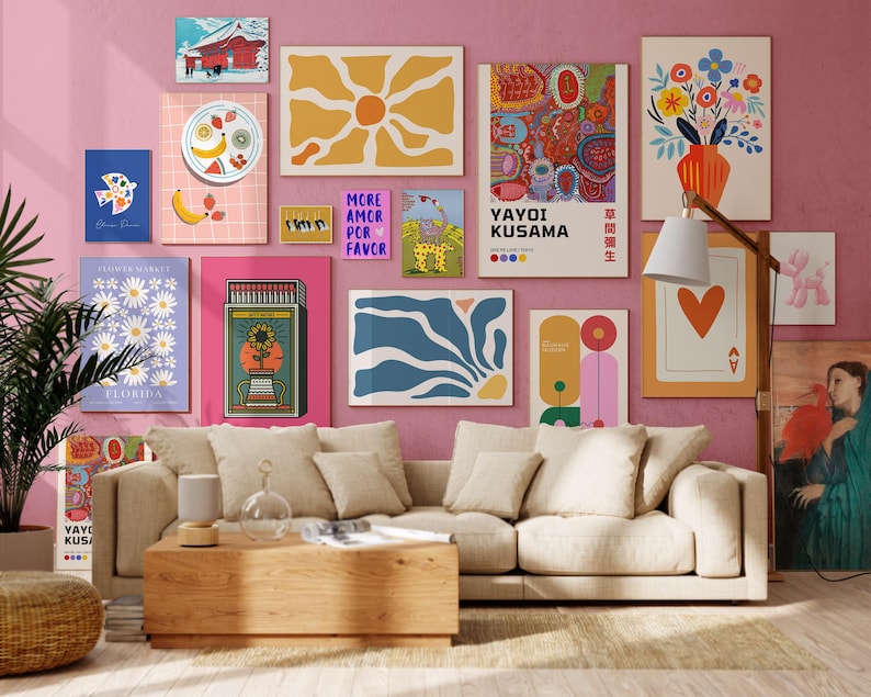 May include: A collection of framed art prints in various sizes and styles, including floral, abstract, and geometric designs. The prints are displayed on a pink wall above a beige sofa and wooden coffee table. A floor lamp and a potted plant are also visible.