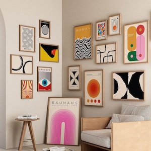 May include: A collection of framed Bauhaus art prints in various sizes and designs, mounted on a white wall. The prints feature geometric shapes, bold colors, and the word "Bauhaus". A wooden chair and side table are in the foreground.