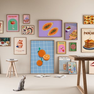 May include: A collection of framed art prints featuring colorful illustrations of food and drinks. The prints include images of cocktails, fruit, and pancakes. The art is displayed on a light-colored wall, creating a vibrant and inviting atmosphere.