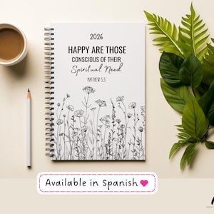 May include: A white spiral-bound 2026 planner with the text "HAPPY ARE THOSE CONSCIOUS OF THEIR Spiritual Need" and "MATTHEW 5:3". The cover features a floral line art design. A cup of coffee, a pencil, and greenery are also present.
