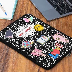 May include: Black laptop sleeve with a composition book design and colorful sticker embellishments. Features a smiley face, a pink purse, a graduation cap, and a pencil. The name "Mrs. Smith" is written on the sleeve.