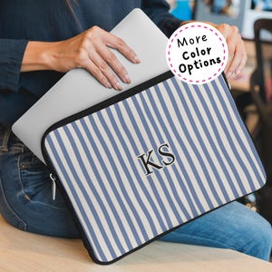May include: A laptop case with a blue and white striped pattern. The case has the initials "KS" in black. The words "More Color Options" are in a pink circle. The case is held by a person wearing a navy blue shirt and blue jeans.