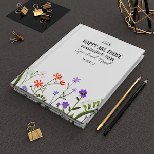 May include: A white 2026 journal with floral illustrations and the text "HAPPY ARE THOSE CONSCIOUS OF THEIR Spiritual Need" and "MATTHEW 5:3". Gold and black pencils and gold binder clips are also visible.