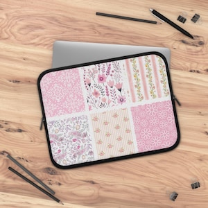 May include: A pink and white floral laptop sleeve with a black zipper and trim. The sleeve features a patchwork design with various floral patterns, including daisies, stripes, and botanical prints. The sleeve is resting on a wooden surface.