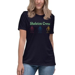 May include: A navy blue t-shirt with the text "Skeleton Crew" in green. Three skeletons are depicted in red, blue, and yellow.