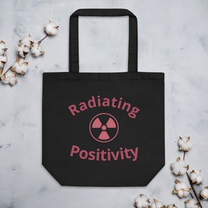 May include: Black tote bag with a pink radiation symbol and the text "Radiating Positivity" printed on it.