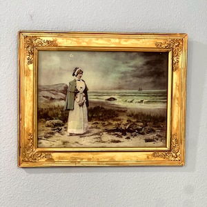 May include: A framed print featuring a person in a long white dress and a green cape standing on a sandy beach near the ocean. The scene includes a rocky shoreline, a dark cloudy sky, and a distant sailboat. The frame is ornate, gold, and metallic.