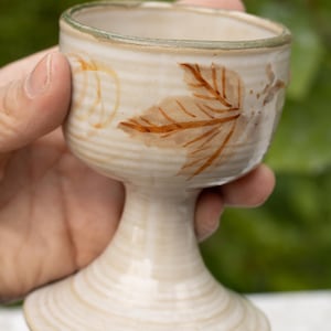 May include: A handmade ceramic goblet with a beige glaze and a leaf design in brown. The goblet has a stem and a wide, shallow bowl. The rim is a darker shade of beige. The goblet is held in a hand.