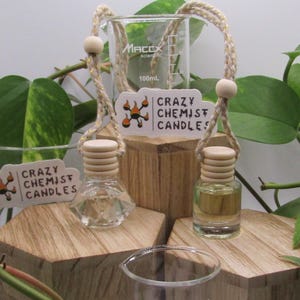 May include: Two glass bottles with wooden caps and gold string hanging from them. The bottles are filled with clear liquid. The bottles are sitting on a wooden surface. The bottles have a label that says "Crazy Chemist Candles".