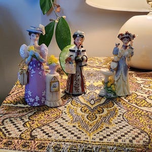 Könnte beinhalten: Drei Porzellanfiguren in historischer Kleidung. Eine trägt ein violettes Kleid mit weißen Blumen, eine andere einen weißen Mantel mit roten Mustern und die dritte ein blau-cremefarbenes Kleid mit einem Vogel neben einem Vogelbad.