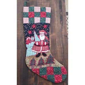 May include: A quilted Christmas stocking featuring a patchwork cuff and toe in red and green with gold stars and moons. The center panel is dark blue with stars, a central Santa Claus figure, and a gold base with three green pine tree appliques.