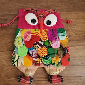 May include: A handmade owl-shaped drawstring bag. The bag is primarily pink with large white eyes and a red beak. The body is made of colorful fabric scraps in various patterns and colors, including red, yellow, green, and orange. The owl has plaid feet and a drawstring closure.