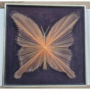 May include: A framed string art piece featuring a butterfly design created with thin, copper-colored metallic threads stretched between small nails. The butterfly is set against a dark purple velvet background within a thin silver-toned metal frame.
