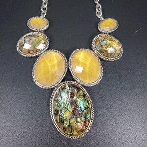 May include: A statement necklace featuring five oval-shaped cabochons set in silver-toned rope-textured metal frames. The design includes two iridescent abalone shell stones and three smooth, translucent yellow stones, all suspended from a silver chain.