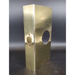 May include: A gold-colored metal door plate with a rectangular shape and a curved edge. It features a keyhole and a circular opening, along with two smaller holes. The plate has a shiny, reflective surface.