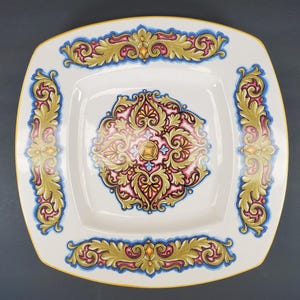 May include: Square white ceramic plate with ornate floral designs in gold, pink, blue, and yellow. The plate features a central medallion and decorative borders, with a yellow rim. This decorative plate is suitable for serving food or as a decorative piece.