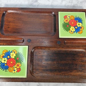 May include: A vintage wooden serving tray with two recessed areas and two square green tiles. Each tile features a colorful floral design with red, yellow, blue, and green flowers. The tray is a dark brown color.