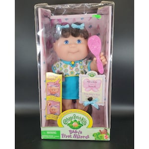 May include: A Cabbage Patch Kids Baby's First Haircut doll in a clear plastic box. The doll has brown hair, blue eyes, and is wearing a floral top and blue skirt. It holds a pink comb. The box has text that reads "Haircut Magic!"