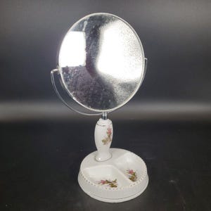 May include: A vintage vanity mirror with a round, silver-toned mirror. The white porcelain base features a small dish with three compartments and floral accents. The mirror is supported by a decorative porcelain stand with pink floral details.