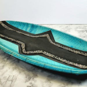 May include: An elongated oval ceramic tray featuring a vibrant turquoise crackle glaze on the outer rim and a matte black center panel. The black section is bordered by a textured, metallic silver strip with a jagged, geometric design.