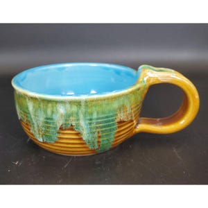 May include: A handmade ceramic soup bowl with a turquoise interior and a textured exterior. The exterior features a brown base with green and yellow dripping glaze, and a curved handle. The bowl is designed for serving soups and stews.