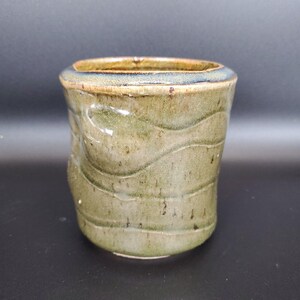 May include: Handmade ceramic tumbler with a unique, textured design. The exterior features horizontal, wave-like patterns in shades of green and brown, with a slightly flared rim. The interior is a darker brown, and the overall aesthetic is rustic and earthy.
