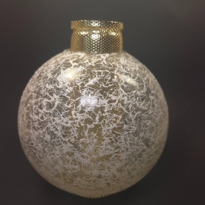 May include: A spherical glass ornament featuring a clear base with a white, web-like, textured pattern across the surface. The top of the ornament is finished with a metallic gold cap that has a fine, perforated, grid-like texture.