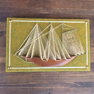 May include: A rectangular wall art piece featuring a wooden ship hull mounted on a mustard yellow felt background. The ship's sails are created using beige string threaded between metal pins, framed by thin gold-toned metal rods at the edges.