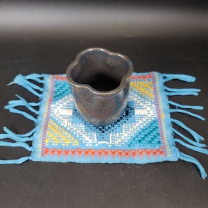 May include: A small, dark blue ceramic cup with a unique, scalloped rim sits on a square, woven coaster. The coaster features a geometric pattern in shades of blue, white, yellow, and orange, with a fringed edge.