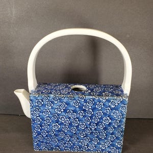 May include: A square blue and white floral patterned teapot with a white handle and spout. The teapot has a rectangular shape with a white handle that arches over the top. The floral pattern is a repeating design of small white flowers on a blue background.