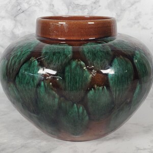 May include: A round ceramic vase with a wide body and a short, straight neck. The surface features a glossy glaze with a deep brown base color overlaid with a textured, mottled pattern of dark emerald green brushstrokes throughout the exterior.