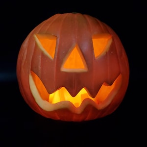 May include: A carved orange pumpkin with triangular eyes and a zig-zag mouth, illuminated from within by a warm, orange glow. The Halloween decoration is set against a black background.