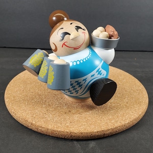 May include: A wooden figurine of a smiling woman in a blue dress, holding two mugs and a small tray with food. The figurine has a brown bun and black shoes. The base is a round cork coaster.