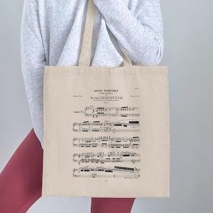 May include: A beige tote bag displaying sheet music from Beethoven's Sonata Pathétique. The text "SONATE PATHETIQUE" is printed at the top, with musical notes below. The bag has long, beige handles.