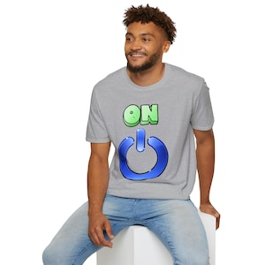 Puede incluir: Una camiseta gris con la palabra "ON" en verde y un gráfico de botón de encendido azul. La camiseta la lleva una persona con vaqueros azules.