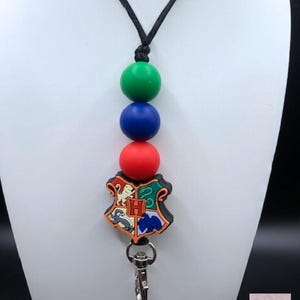 May include: A lanyard featuring a colorful crest with a silver clasp. The lanyard has three large, round beads in green, blue, and red. The crest is a shield shape with a black border and various colorful images.