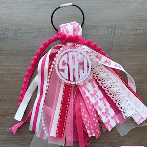 May include: A keychain tassel featuring a black metal ring and a collection of pink and white ribbons, including lace, pom-pom trim, and sheer fabric. A circular clear acrylic charm is attached, displaying the white monogram letters SAJ in a script font.