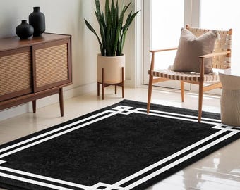 May include: A rectangular black rug with a white geometric border. The rug is placed on a light-colored floor in a room with a wooden cabinet, a chair, and a potted plant. The rug's dimensions are approximately 60 inches by 96 inches.