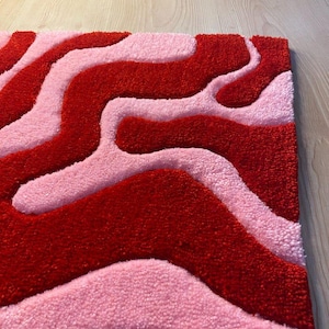 May include: A rectangular rug featuring a wavy design in red and pink hues. The rug has a soft, textured pile, with the red and pink sections forming a contrasting pattern. It is placed on a light-coloured wooden floor.