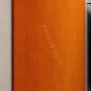 May include: An orange rectangular rug with a textured surface. The rug is angled against a white wall. The text "the royal rug co" is printed on the rug.