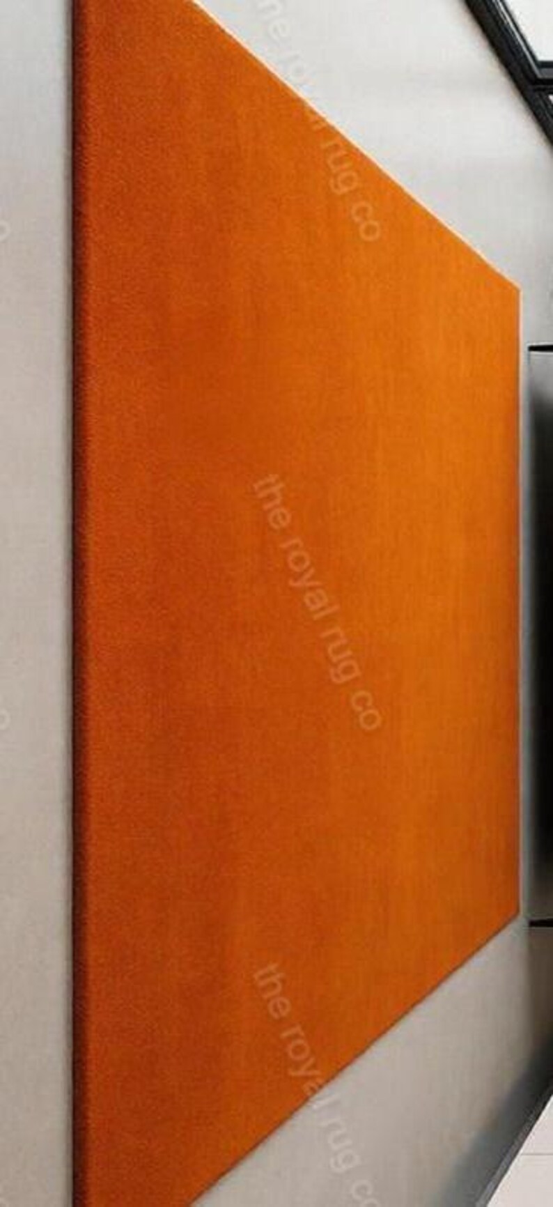 May include: An orange rectangular rug with a textured surface. The rug is angled against a white wall. The text "the royal rug co" is printed on the rug.