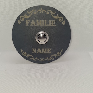 Könnte beinhalten: Eine schwarze runde Türklingel mit einem silbernen Knopf. Die Türklingel hat die Worte "Familie" und "NAME" in Gold graviert.