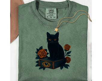 May include: Sage green t-shirt featuring a black cat illustration sitting on a book with an eye symbol. The design includes orange roses and the text "COMFORT COLORS". A gold star pendant necklace is draped across the shirt.