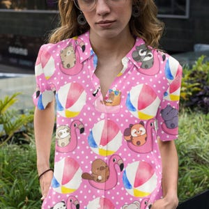 Puede incluir: Camisa rosa con botones con un estampado de animales de dibujos animados en flotadores inflables y pelotas de playa. La camisa tiene mangas cortas y un estampado de lunares blancos. Los animales incluyen perezosos, mapaches e hipopótamos.