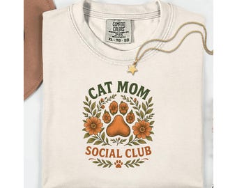 May include: Cream-colored t-shirt with the text "CAT MOM SOCIAL CLUB" in a circular design featuring a cat paw print and orange flowers. A gold necklace with a star pendant rests on the shirt. The shirt is folded and has a Comfort Colors tag.